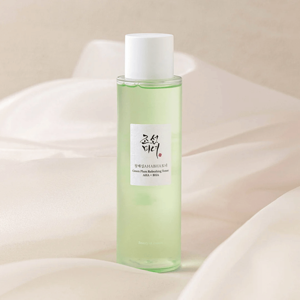 En flaska med "Green Plum Refreshing Toner" AHA + BHA står på en mjuk, ljus bakgrund. Produkten har en grön vätska som avspeglar fräschhet och naturlighet. Etiketten är vit med svarta och röda inslag, vilket ger en elegant och ren look.